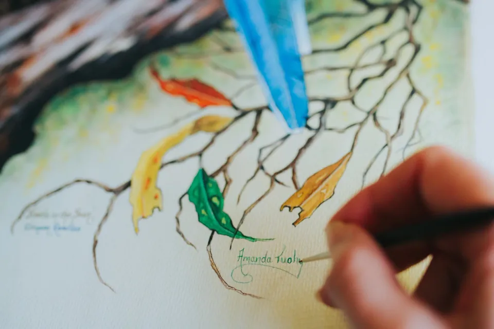 Close-up of Amanda’s hand signing her name in green paint beside autumn gum leaves on one of her paintings.