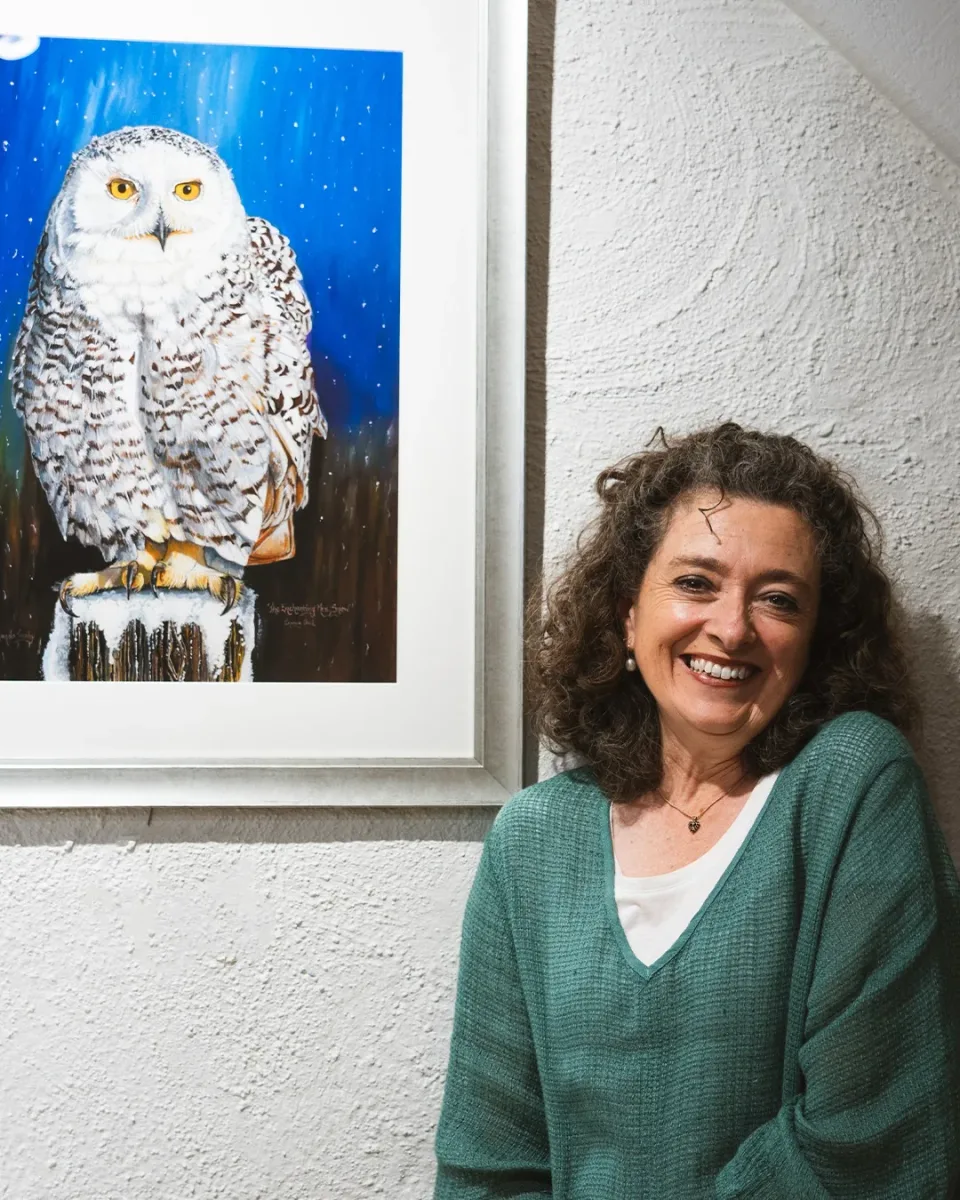 Amanda Tuohy smiling beside a framed painting of a snowy owl, inside Sanctuary Gallery, Hahndorf