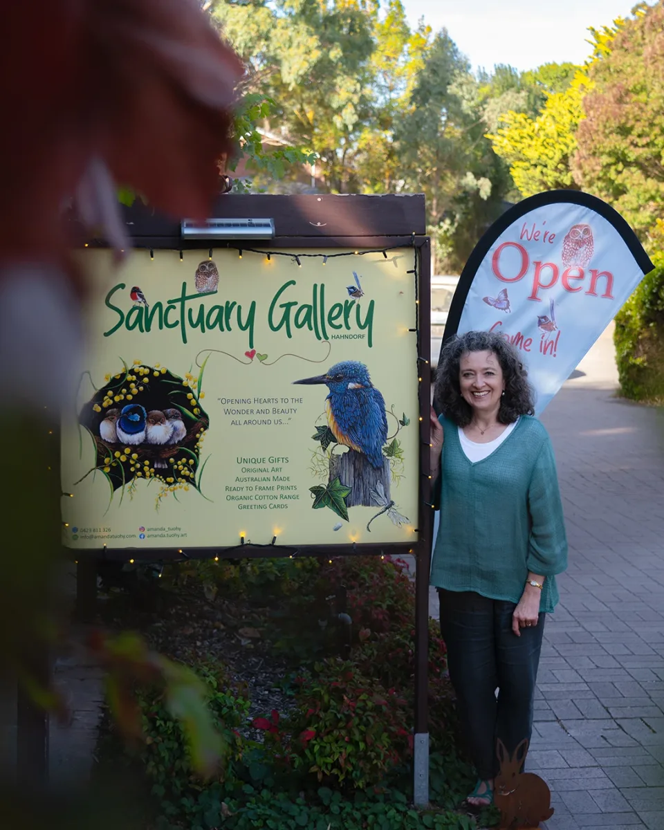 Amanda Tuohy standing beside the Sanctuary Gallery sign and its “We’re Open” flag, outside the gallery in Hahndorf