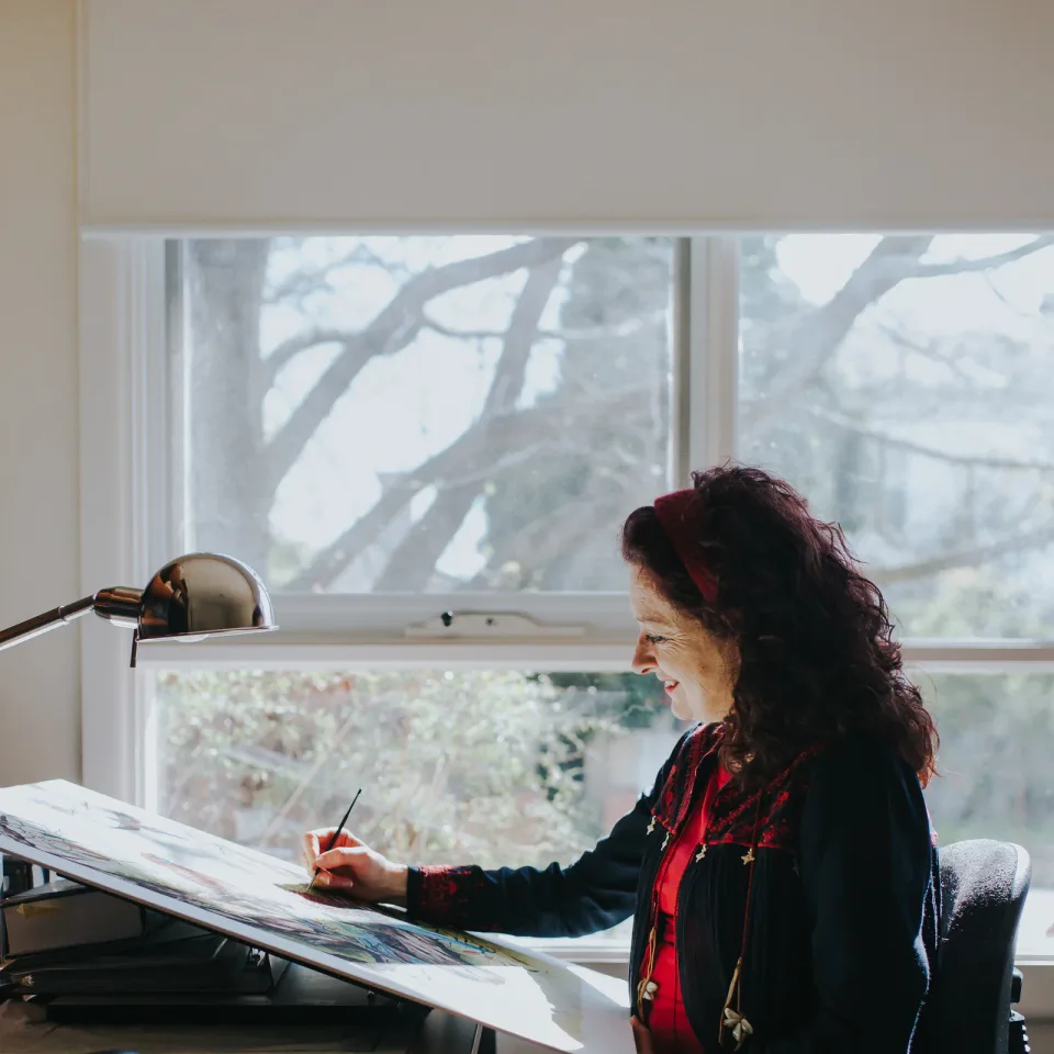 Amanda Tuohy smiles as she paints at a drafting board beside a large window, with bare tree branches outside.
