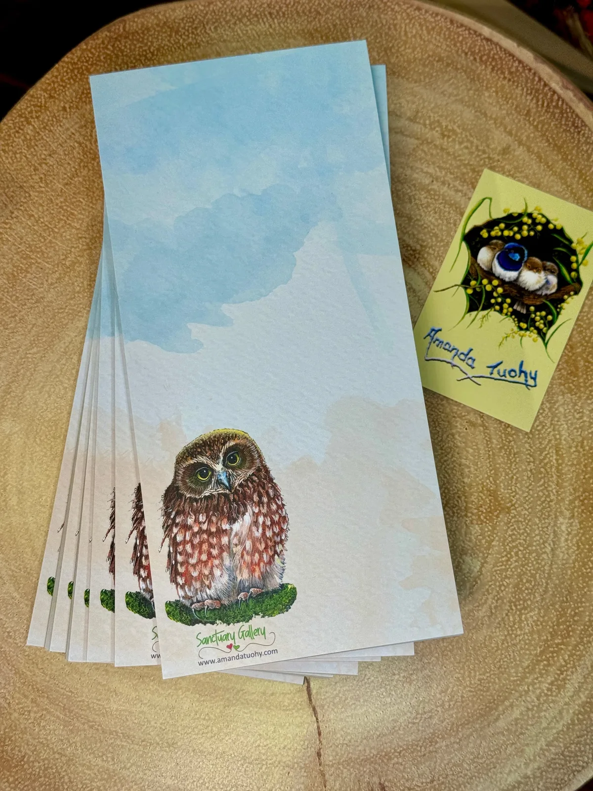 A stack of tall, narrow notepads in pale blue and cream on a round wooden slice, the top one printed with a boobook owl, beside a small yellow card signed Amanda Tuohy.