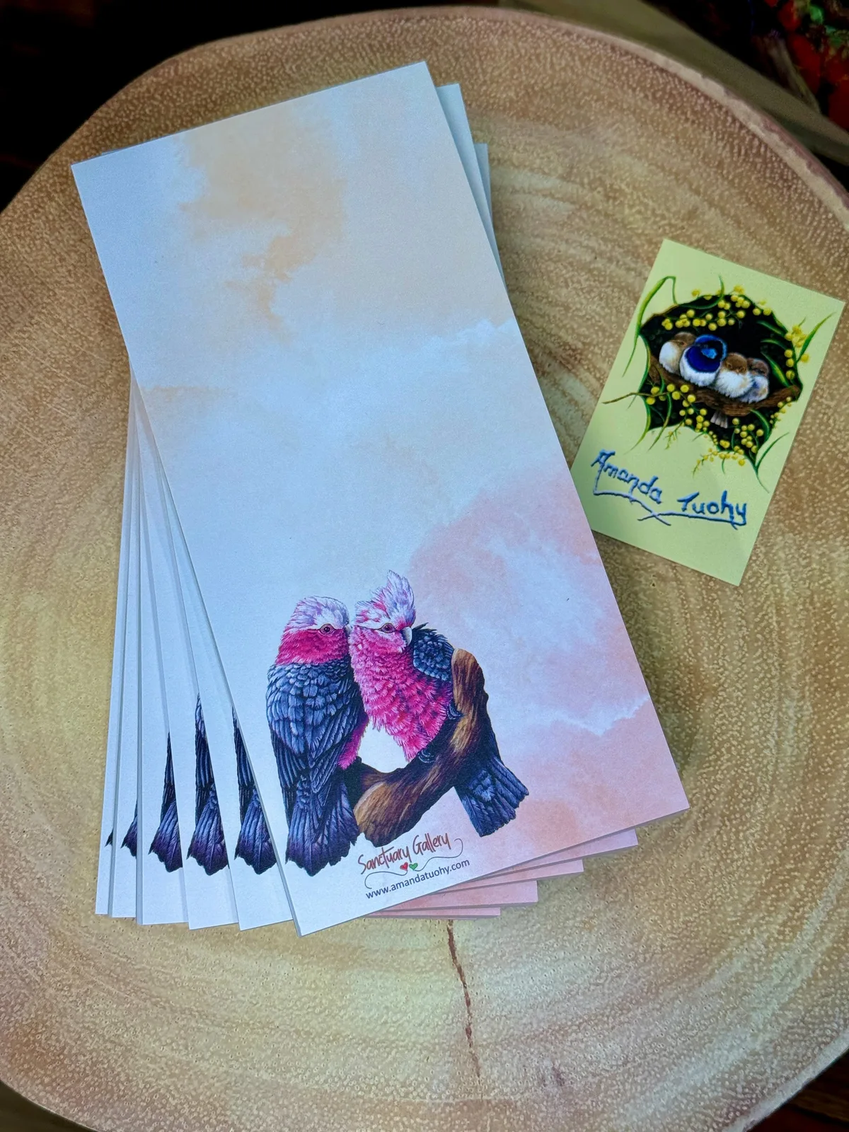 A stack of tall, narrow notepads in pale blue and peach on a round wooden slice, the top one printed with two galahs, beside a small yellow card signed Amanda Tuohy.