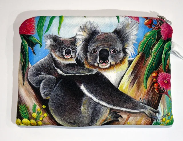 A zip-up cotton pouch printed with a koala carrying a young koala on its back in a gum tree, among pink flowering gum blossoms, green leaves and yellow wattle against a blue sky.