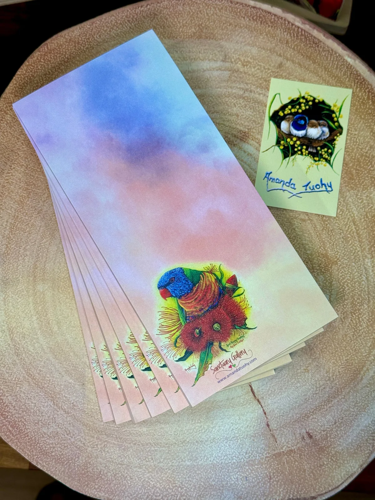 A stack of tall, narrow notepads in lilac, pink and peach on a round wooden slice, the top one printed with a rainbow lorikeet, beside a small yellow card signed Amanda Tuohy.