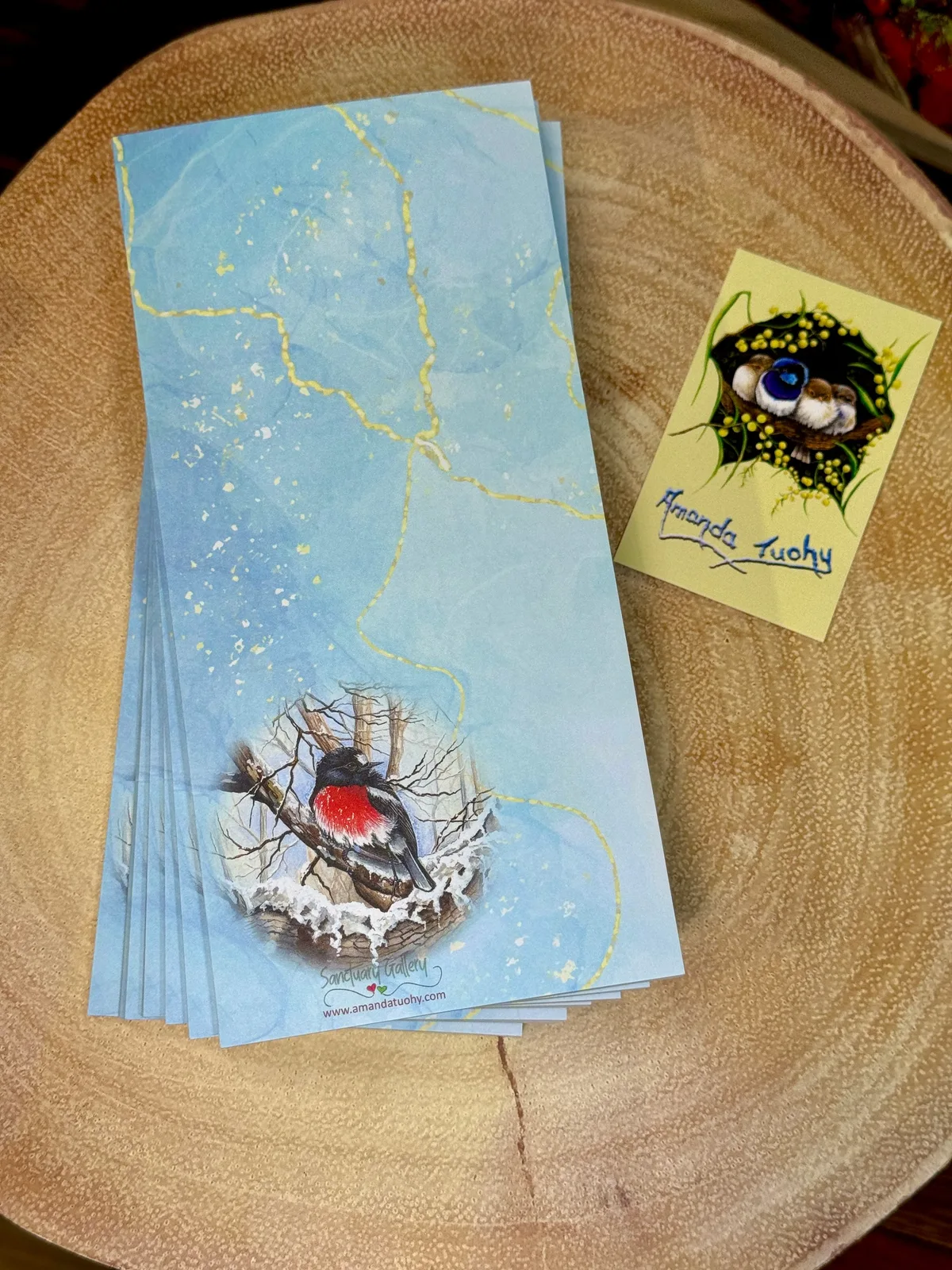 A stack of tall, narrow notepads in pale blue with fine gold lines on a round wooden slice, the top one printed with a scarlet robin on a snow-dusted branch, beside a small yellow card signed Amanda Tuohy.