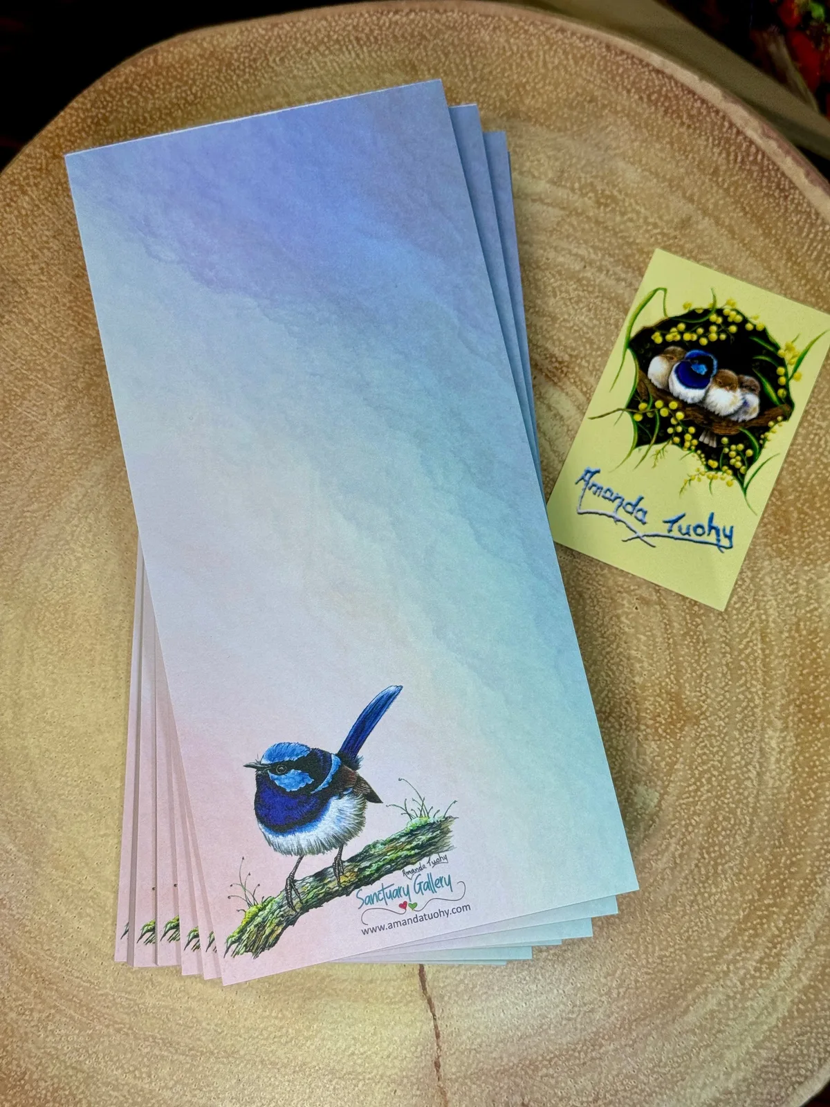 A stack of tall, narrow notepads in blue, lilac and pink on a round wooden slice, the top one printed with a wren, beside a small yellow card signed Amanda Tuohy.