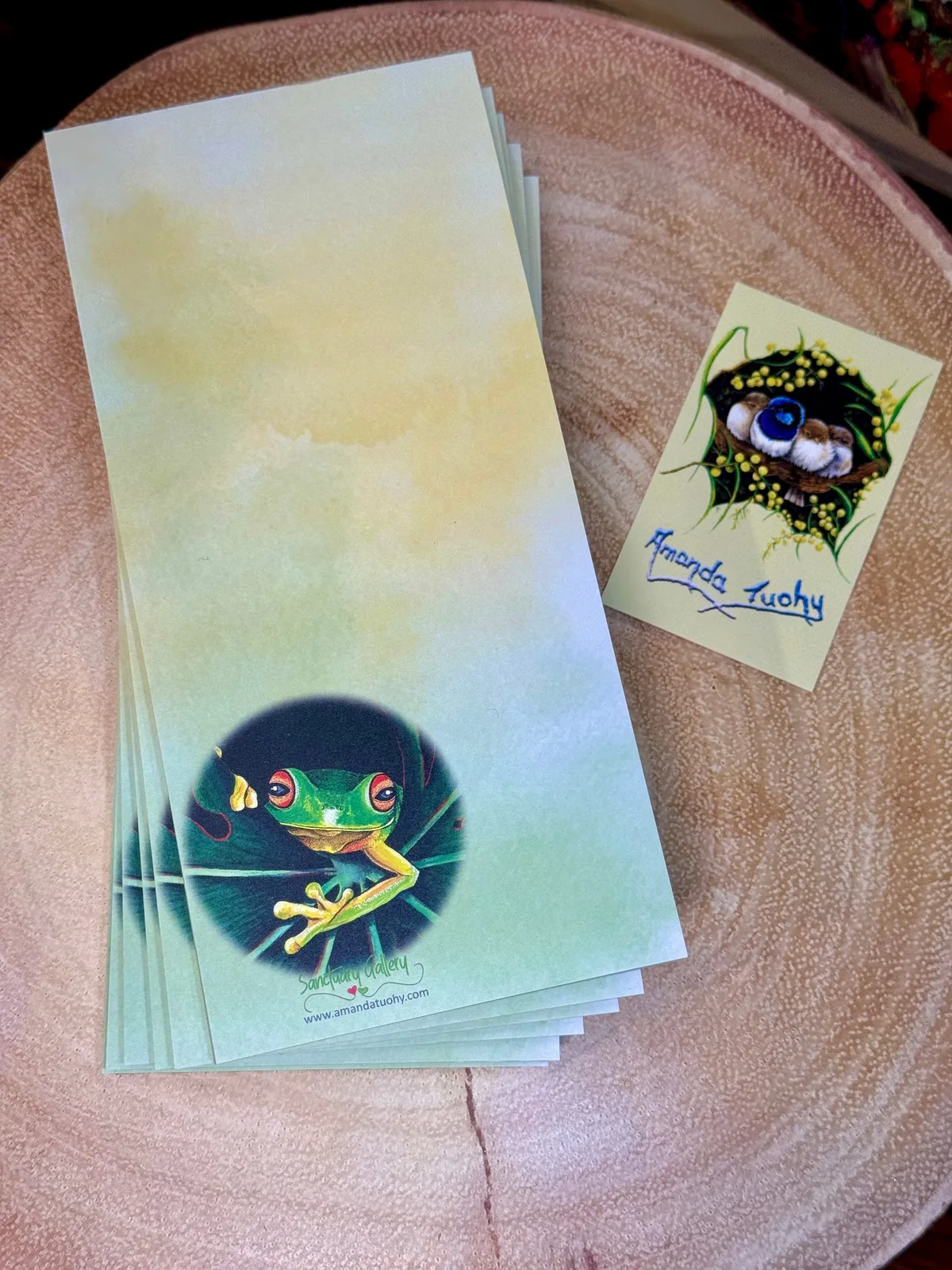 A stack of tall, narrow notepads in pale green and yellow on a round wooden slice, the top one printed with a red-eyed tree frog, beside a small yellow card signed Amanda Tuohy.