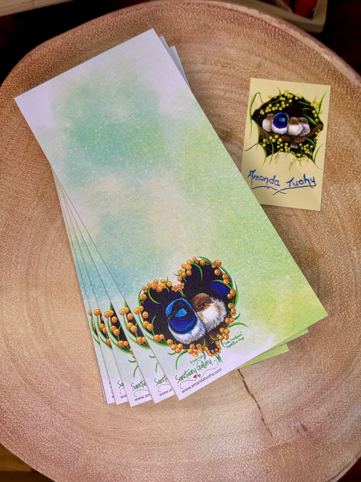 A stack of tall, narrow notepads in soft green on a round wooden slice, the top one printed with two small wrens in a heart of golden wattle, beside a small yellow card signed Amanda Tuohy.
