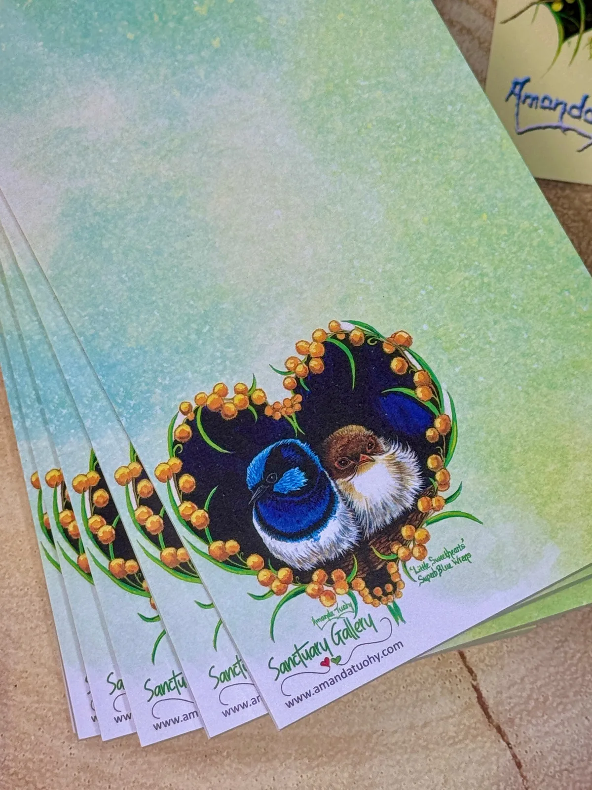 A pad of notepaper washed in blue-green and yellow, printed with two small wrens, one bright blue and black and one soft brown, nestled together in a heart of golden wattle, with the Sanctuary Gallery name and web address beneath.