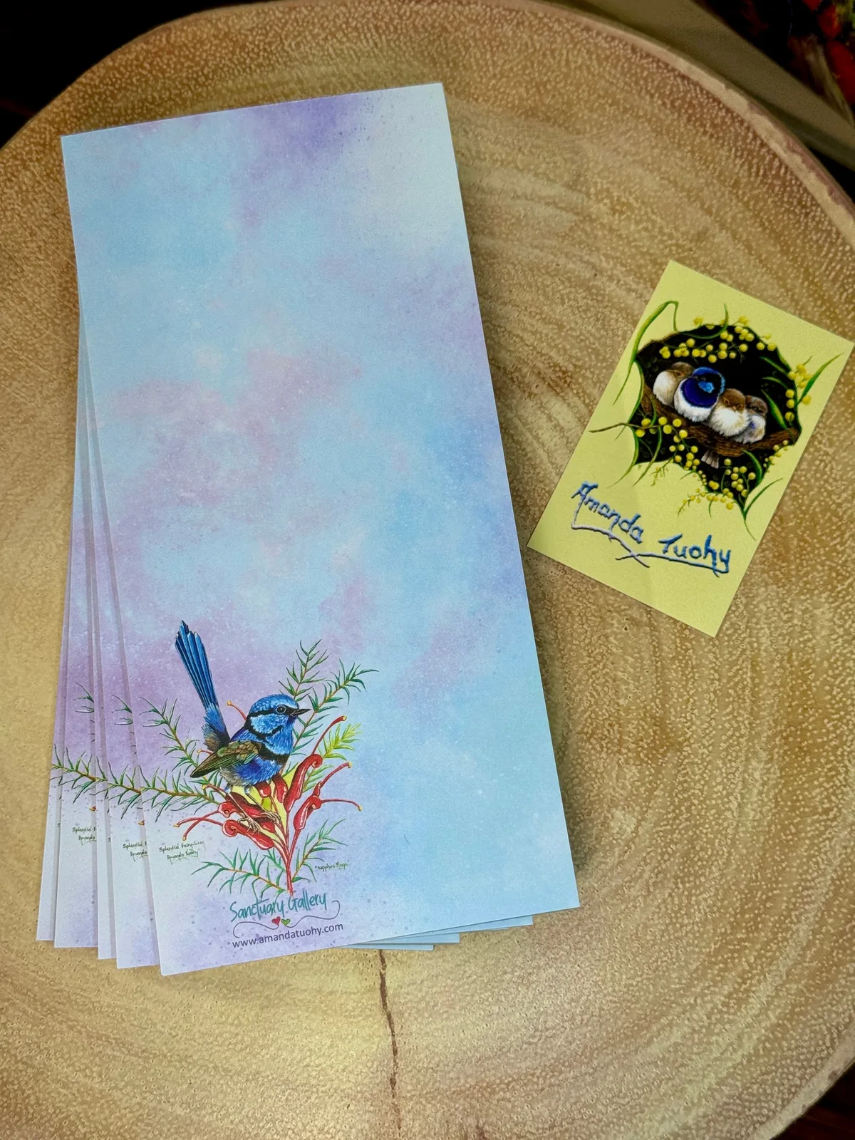 A stack of tall, narrow notepads in blue and lilac on a round wooden slice, the top one printed with a wren on red grevillea flowers, beside a small yellow card signed Amanda Tuohy.